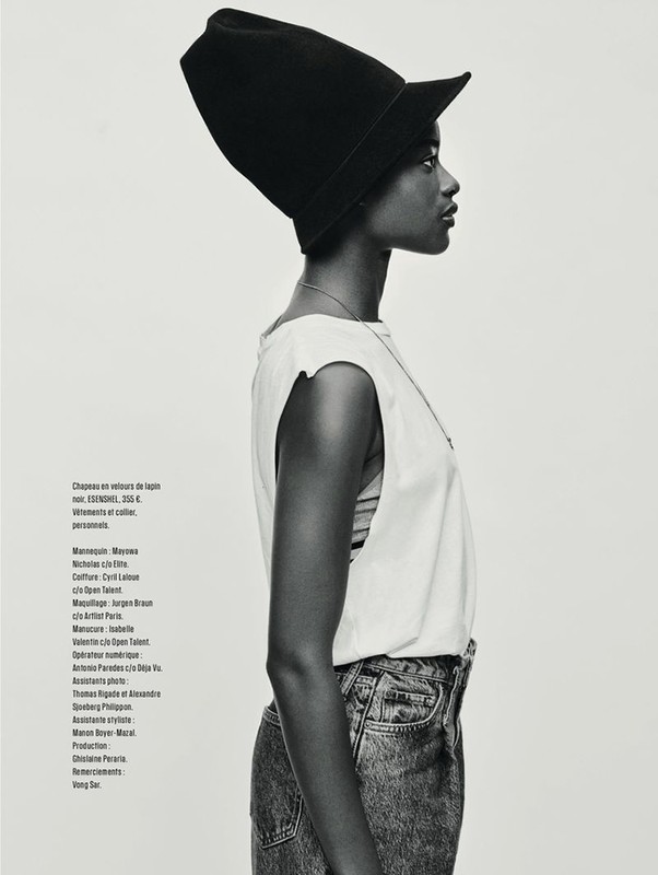 Profile shot of a dark-skinned woman wearing a large black hat, sleeveless top, and jeans against a neutral studio background.