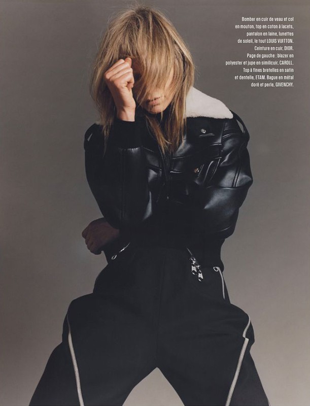 Fashion editorial portrait of a female model in a black leather jacket and track pants against a solid gray studio backdrop.