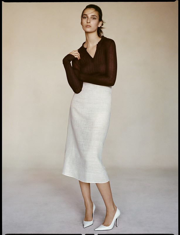Elegant brunette poses in photo studio wearing a brown sheer top, white midi skirt, and white high heels against a neutral backdrop.