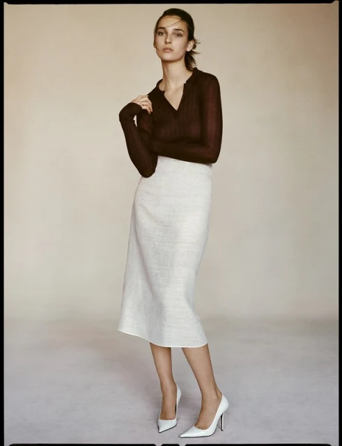 Elegant brunette poses in photo studio wearing a brown sheer top, white midi skirt, and white high heels against a neutral backdrop.
