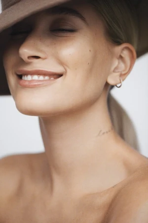 Close-up portrait of Hailey Bieber smiling with eyes closed, wearing a wide-brim hat and a small hoop earring against a neutral background.