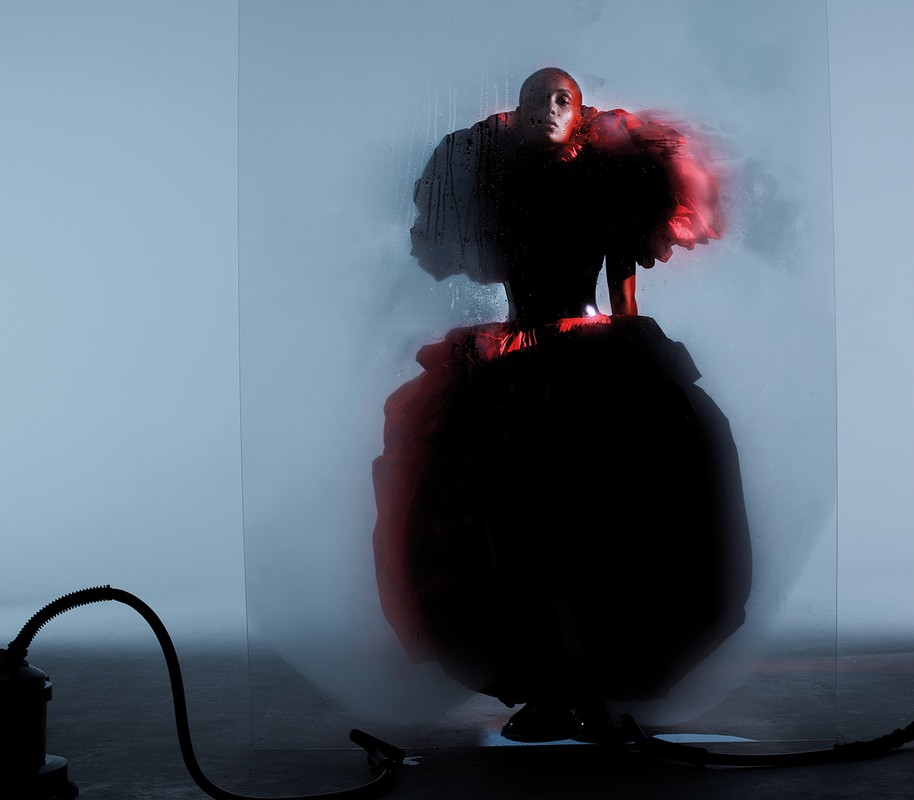 Person in elaborate black dress posing behind frosted glass with studio equipment, highlighted by red and blue lighting.