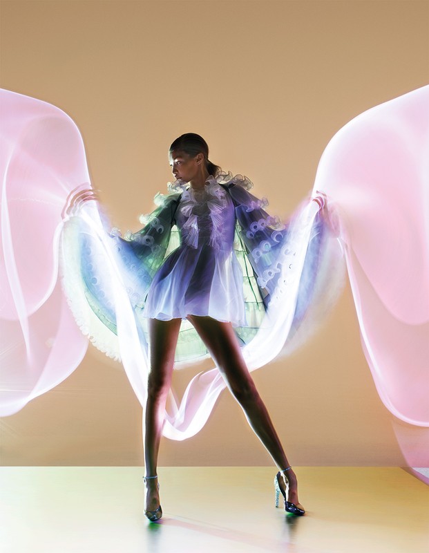 A stylish woman in a short blue dress stands in a photo studio, holding up sheer pink fabric, showcasing fashion and photographic techniques.
