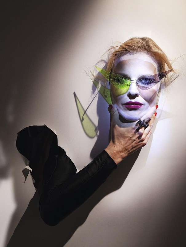 Woman wearing sunglasses with geometric color filters, hand holding head against a white wall with a shadow, red nail polish.
