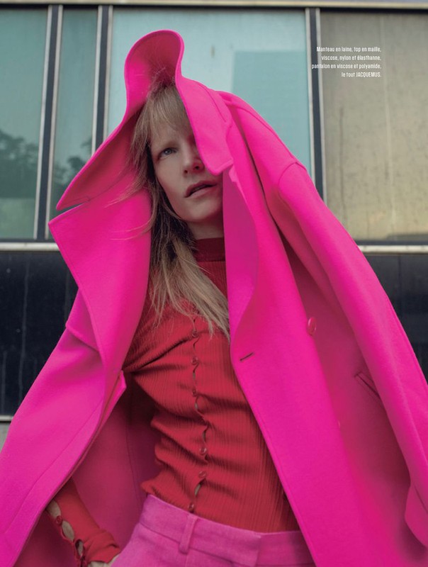 A woman in a vibrant pink coat and matching pants stands outdoors against a modern building with glass and metal accents.