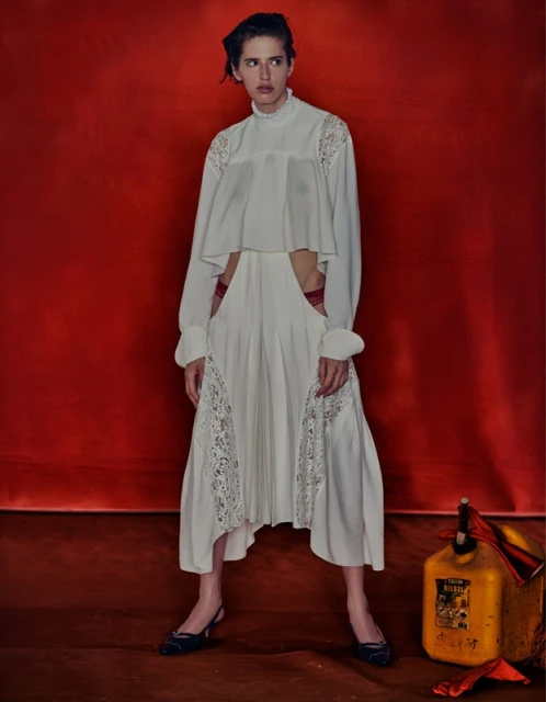 A young woman in a long-sleeved white gown poses indoors against a red backdrop with a vintage yellow gas container.