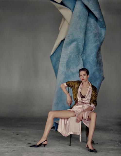 Fashion shot of a woman in a pink satin robe and green jacket sitting on a chair against a gray backdrop with abstract blue fabric.
