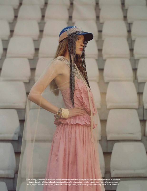 Fashion editorial shot of a long-haired woman in a pink dress and blue cap posing against a stadium seating backdrop.