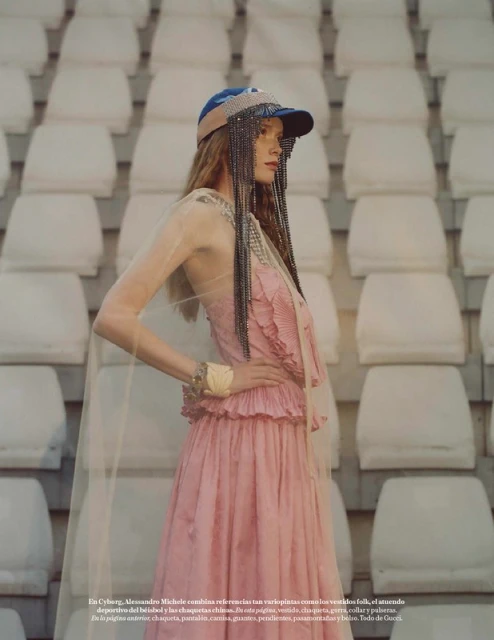 Fashion editorial shot of a long-haired woman in a pink dress and blue cap posing against a stadium seating backdrop.