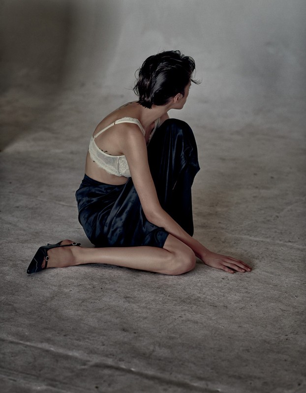 Woman with short dark hair poses kneeling in a cream bra and navy skirt in a photo studio with a blurred background and grey floor.