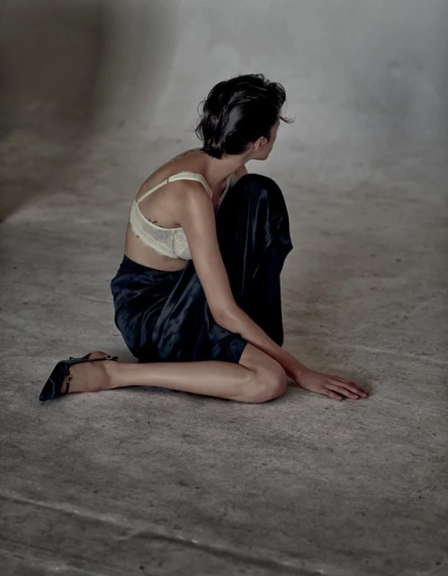 Woman with short dark hair poses kneeling in a cream bra and navy skirt in a photo studio with a blurred background and grey floor.