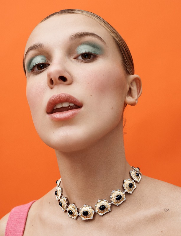 Close-up shot of a young woman with distinctive makeup and a vintage necklace, set against a bright orange backdrop.