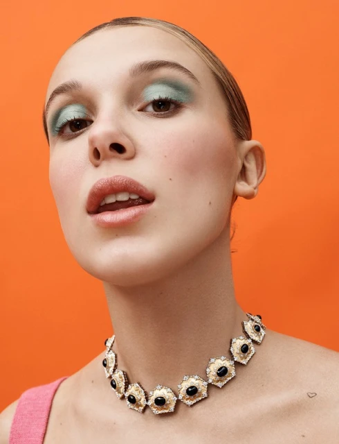 Close-up shot of a young woman with distinctive makeup and a vintage necklace, set against a bright orange backdrop.
