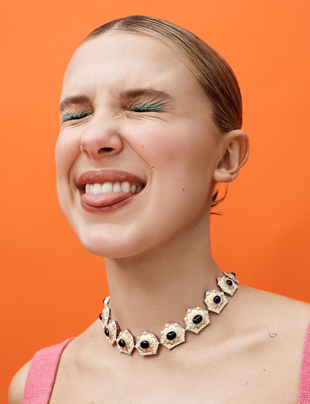 Close-up shot of a happy young adult woman with eyes closed, sticking out tongue, and wearing a gemstone necklace against orange backdrop.