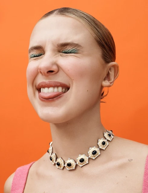 Close-up shot of a happy young adult woman with eyes closed, sticking out tongue, and wearing a gemstone necklace against orange backdrop.