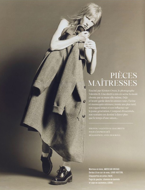 Editorial fashion shot of a blonde woman in a gray coat, black shoes, and white socks against a neutral background.