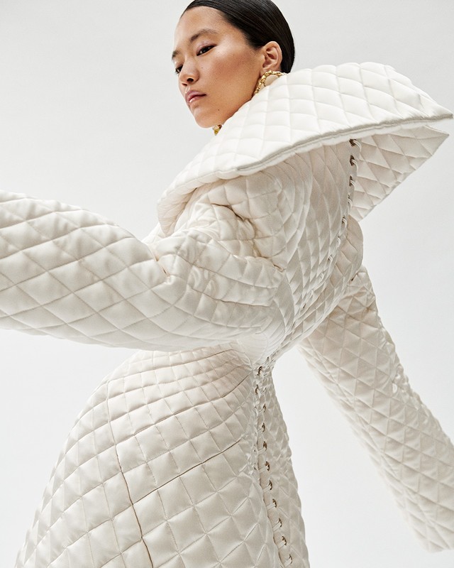 Elegant Asian woman poses in a striking white quilted coat with gold earrings against a clean white backdrop in a fashion setting.