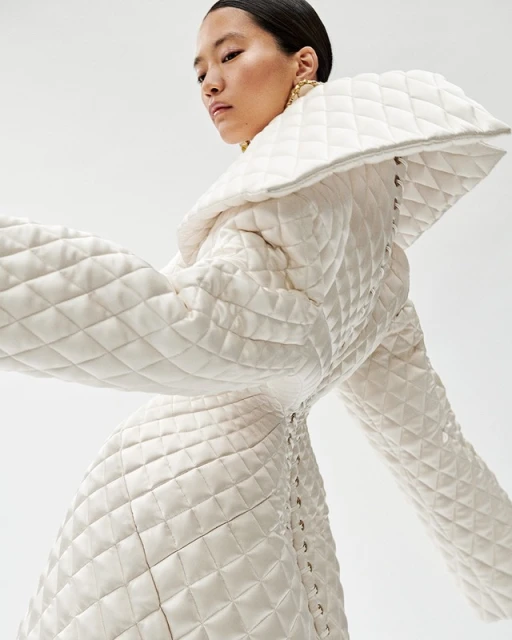 Elegant Asian woman poses in a striking white quilted coat with gold earrings against a clean white backdrop in a fashion setting.
