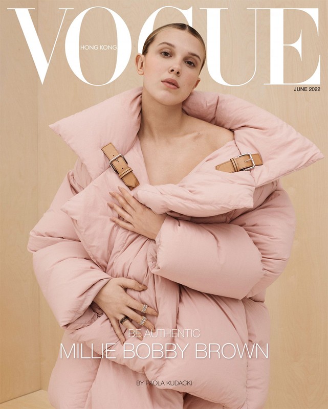 Millie Bobby Brown wearing a voluminous pink puffer jacket with tan leather buckle straps on a wood-paneled background.