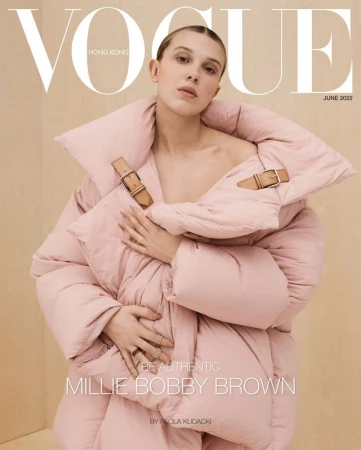 Millie Bobby Brown wearing a voluminous pink puffer jacket with tan leather buckle straps on a wood-paneled background.