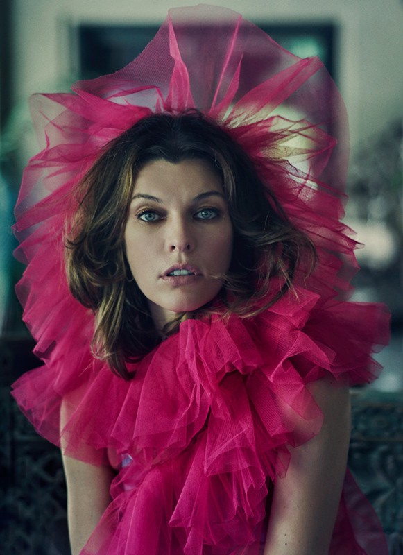 Close-up portrait of a woman with blue eyes wearing a large pink tulle ruffled collar. Indoor setting with blurred background.