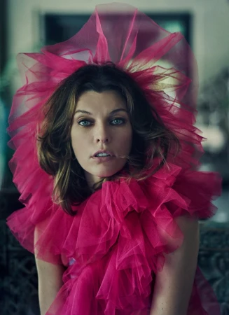 Close-up portrait of a woman with blue eyes wearing a large pink tulle ruffled collar. Indoor setting with blurred background.