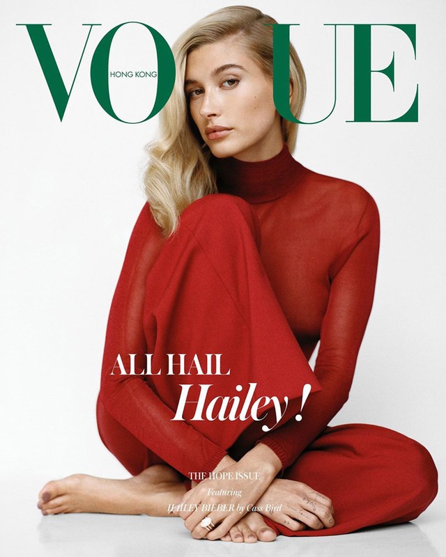 Hailey Bieber sitting barefoot in a sheer red turtleneck top and matching trousers against a white background for Vogue Hong Kong.