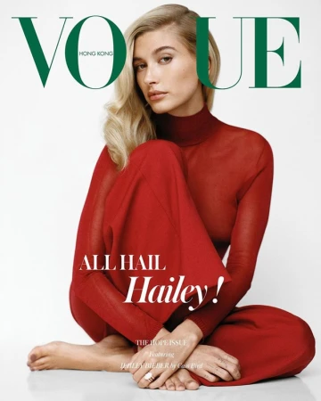 Hailey Bieber sitting barefoot in a sheer red turtleneck top and matching trousers against a white background for Vogue Hong Kong.
