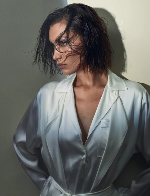 Close-up of a woman with wet, short dark hair, wearing a silky white robe, looking off to the side in a softly lit, indoor setting.