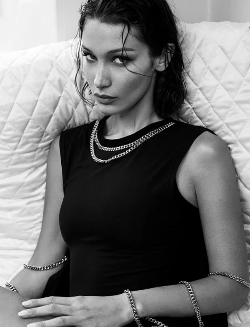 Black and white portrait of a model wearing a black sleeveless top and layered silver chain necklaces, against a quilted background.