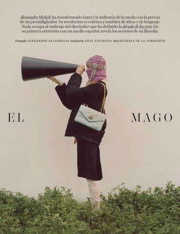 Fashion editorial shot: A model in a black coat and embellished mask holds a large megaphone in front of a neutral background with greenery.