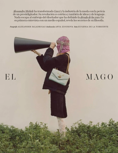 Fashion editorial shot: A model in a black coat and embellished mask holds a large megaphone in front of a neutral background with greenery.