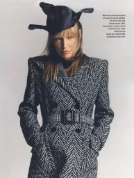 Model wearing a black and white herringbone coat with a wide belt and a sculptural black hat against a grey background