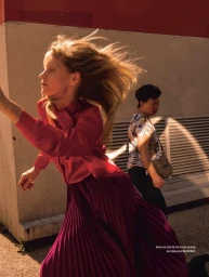 Blonde woman in vibrant pink silk blouse and purple pleated midi skirt walking outdoors with hair blowing in wind