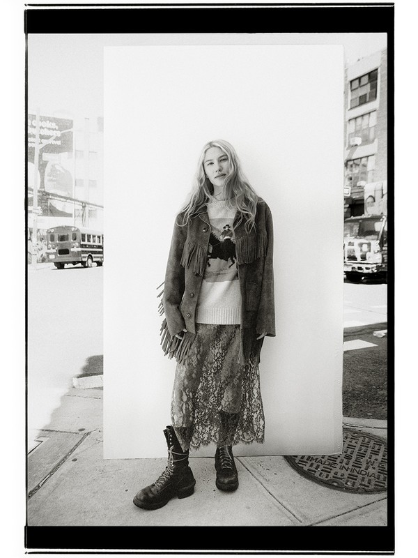 Black-and-white portrait of a woman in an outdoor urban setting, wearing a patterned skirt, boots, and a fringed jacket against a white wall.