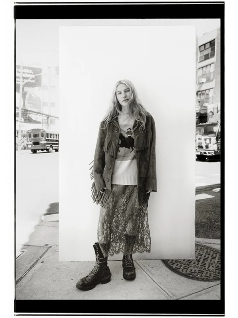 Black-and-white portrait of a woman in an outdoor urban setting, wearing a patterned skirt, boots, and a fringed jacket against a white wall.