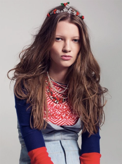 Young woman with long wavy hair wearing a tiara, red top and necklaces poses in a studio setting against a neutral backdrop.