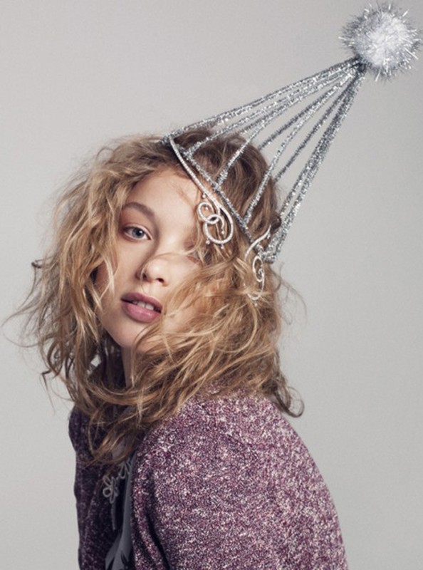 A young adult female poses indoors with a silver glittery party hat against a neutral background, wearing a maroon knit sweater.
