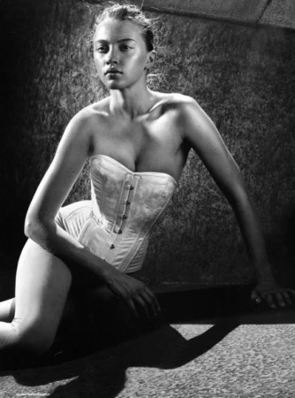 Model wearing a white strapless corset posing against a textured dark surface in a high-contrast black and white portrait.