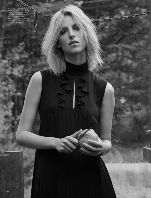 Black-and-white portrait of a blonde adult female in a black dress, holding a citrus fruit outside with a blurry background.
