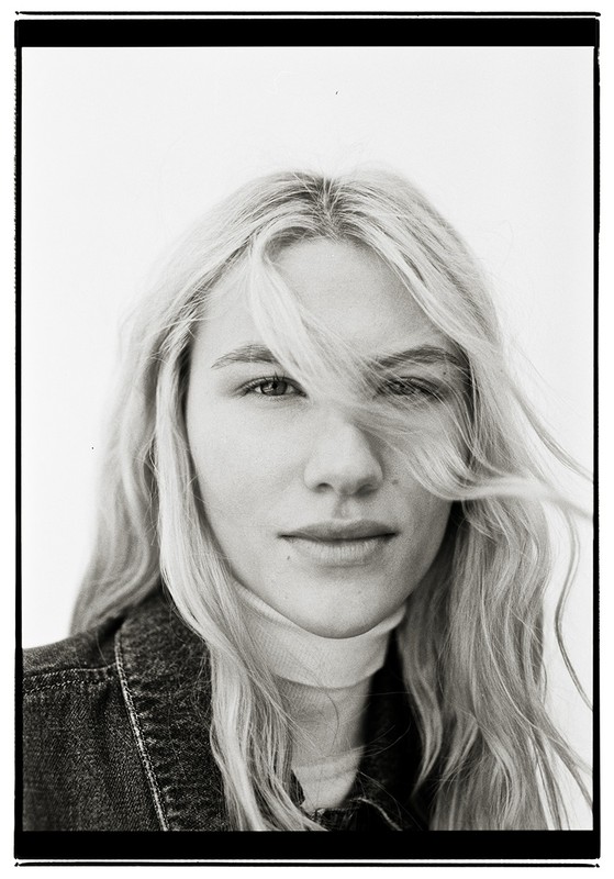 Close-up portrait of a woman with long blonde hair blowing across her face against a plain white background.