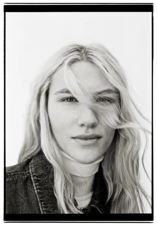 Close-up portrait of a woman with long blonde hair blowing across her face against a plain white background.
