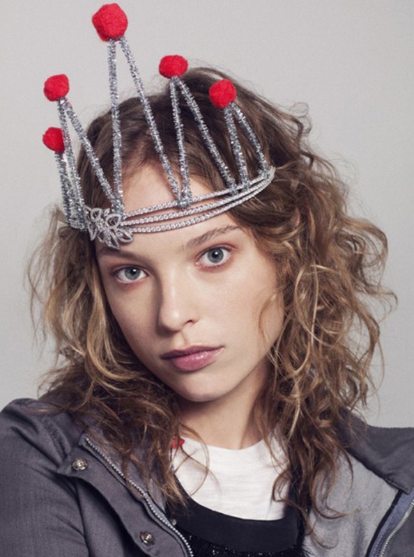 Portrait of a young woman wearing a silver crown with red pom poms, styled with fair skin, wavy blonde hair, and a grey jacket.