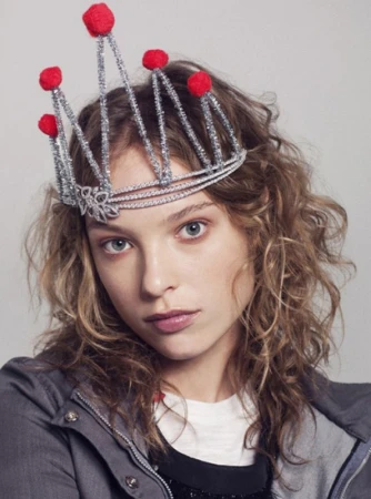 Portrait of a young woman wearing a silver crown with red pom poms, styled with fair skin, wavy blonde hair, and a grey jacket.