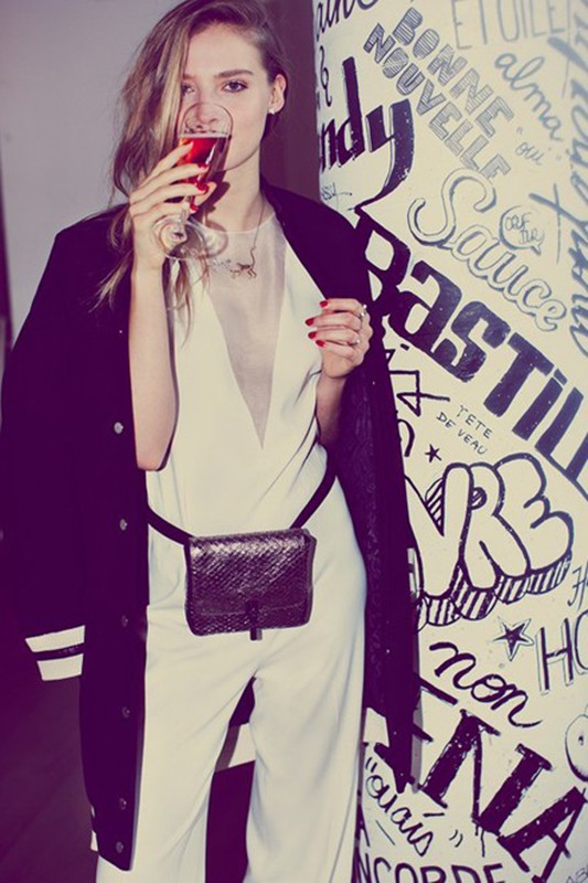 Woman in white jumpsuit and black jacket drinking from a wine glass in front of wall with graffiti art, fashion, indoor, studio lighting.