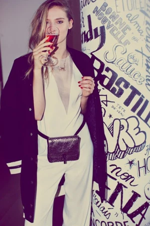 Woman in white jumpsuit and black jacket drinking from a wine glass in front of wall with graffiti art, fashion, indoor, studio lighting.