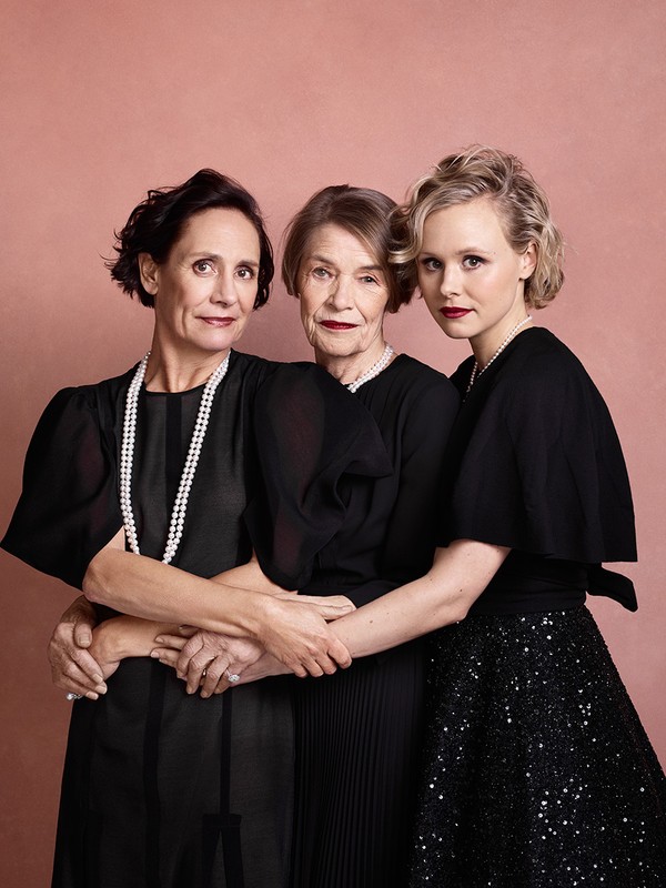 Three women in elegant black dresses and pearl necklaces pose indoors against a neutral pink background in a portrait-style photograph.