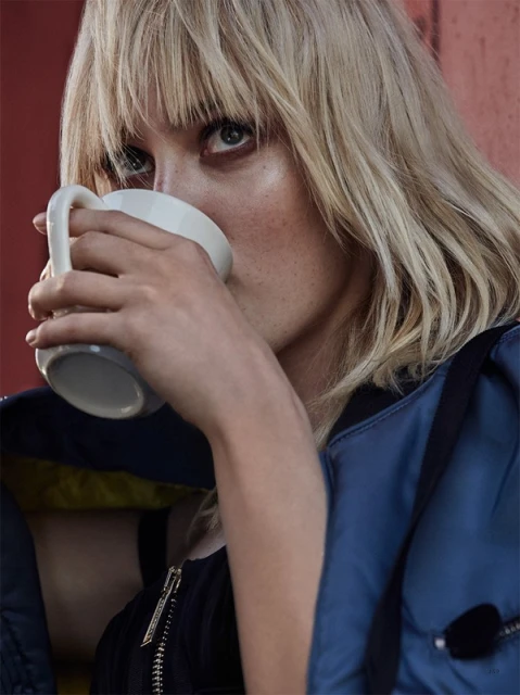 Portrait of a young woman with blond hair drinking from a white mug, wearing a blue jacket with a black shirt, against a solid red wall.