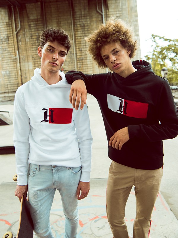 Two young male models wearing graphic hoodies and casual trousers posing outdoors with a skateboard.
