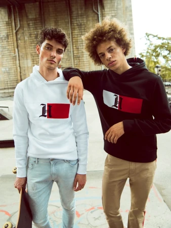Two young male models wearing graphic hoodies and casual trousers posing outdoors with a skateboard.